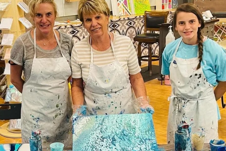 Three women in aprons stand behind a table with blue abstract paintings.