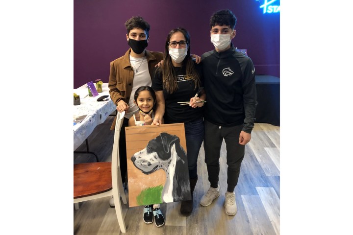 Group of four people, including a child, holding a dog painting indoors, all wearing masks.
