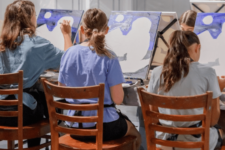Group of people painting at easels in an art class with chairs and colorful artwork on display.