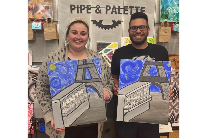 Two people holding paintings of a bridge at a venue with the sign 'Pipe & Palette' in the background.