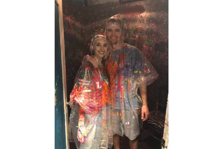 Two people wearing ponchos, covered in colorful paint splatters, smiling indoors.