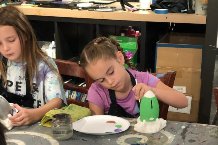 Children painting colorful objects at a table with paints and brushes.