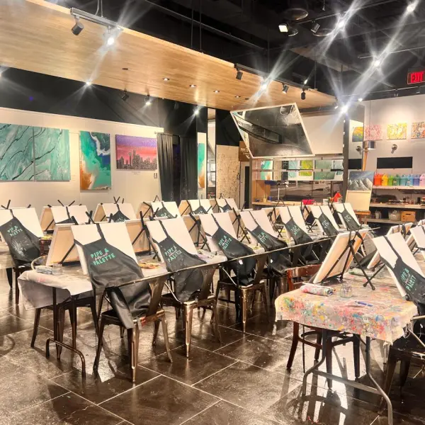 Empty art studio with easels, aprons, and paintings on walls.