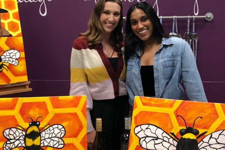 Two women standing with bee-themed art on canvases in a brightly colored room.
