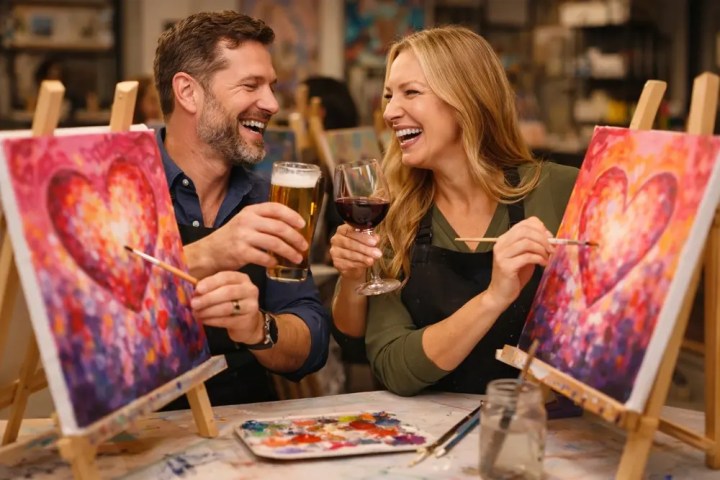 Smiling couple painting heart designs, holding a beer and a wine, in an art class setting.